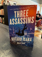 Book titled 'Three Assassins' by Kotaro Isaka on a surface with blurred background