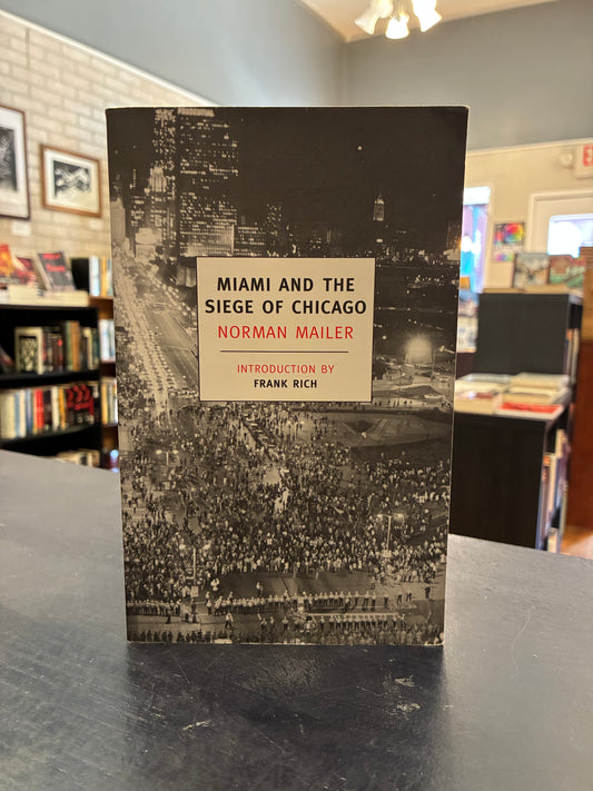  'Miami and the Siege of Chicago' by Norman Mailer, standing upright on a dark tabletop in a bookstore or library interior.