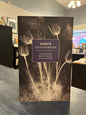 book titled 'Ghosts' by Edith Wharton, standing upright on a dark tabletop in a bookstore or library interior. The cover features a dark sepia-toned floral design with a central purple label containing white text.